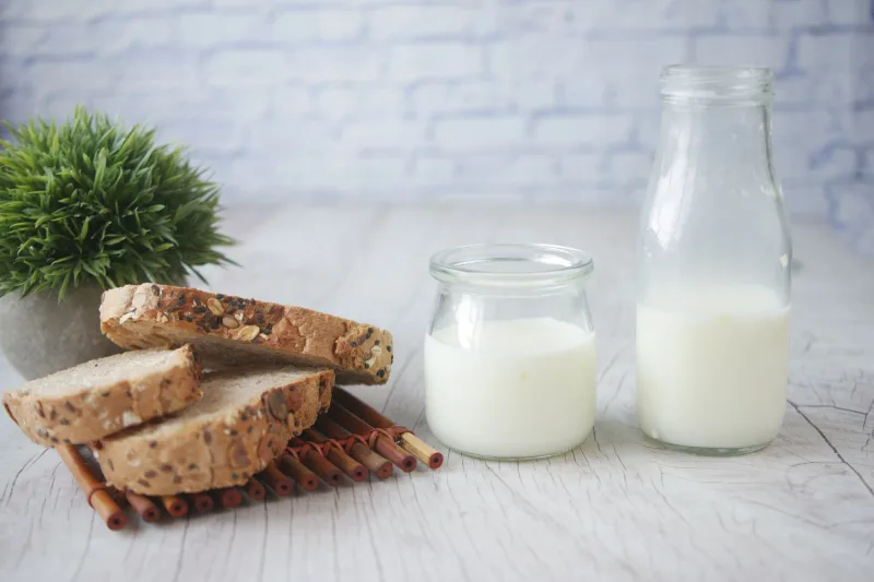 Beyaz ahşap zeminde tahıllı ekmek dilimleri ve cam şişelerde süt